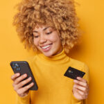 Mulher sorridente segurando cartão de crédito e celular enquanto faz compra online