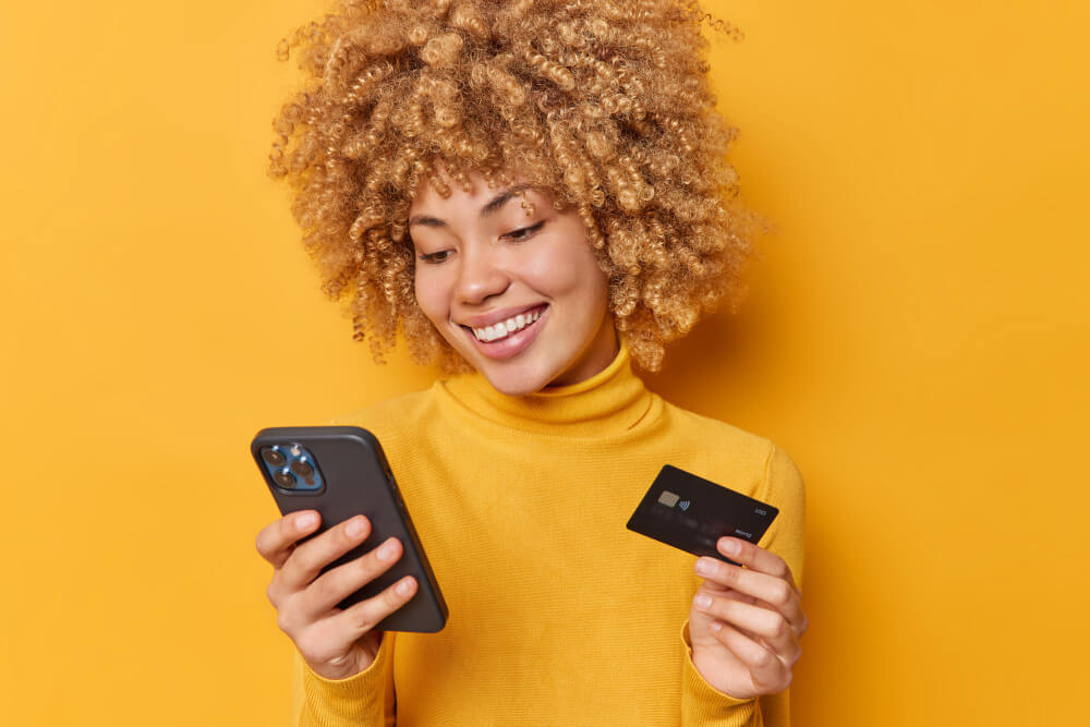 Mulher sorridente segurando cartão de crédito e celular enquanto faz compra online