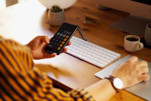 Pessoa usando calculadora no celular em frente ao computador para definir precificação