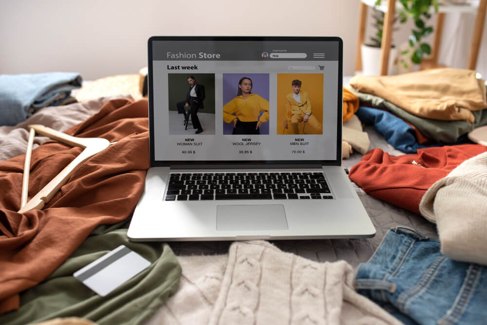 Notebook exibindo loja de moda online sobre cama com roupas e cartão de crédito ao lado