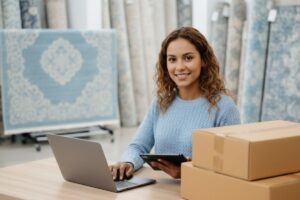 Mulher trabalhando em loja de tapetes com notebook e caixas de envio, representando vendas em marketplaces