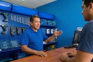 Atendente de meia-idade, vestindo camisa polo azul, gesticulando e explicando sobre ferramentas expostas em um painel azul para um cliente no balcão de uma loja de ferragens.