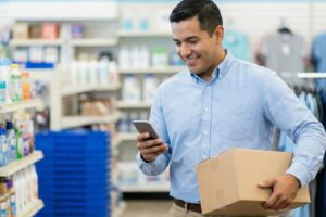 Funcionário em loja segurando caixa enquanto consulta o celular, representando gestão de pedidos e redução de devoluções em marketplaces.