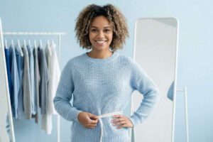 Mulher negra sorrindo, com cabelo cacheado e blusa de tricô azul, segurando uma fita métrica ao redor da cintura em um estúdio de moda com araras de roupas e espelhos ao fundo.