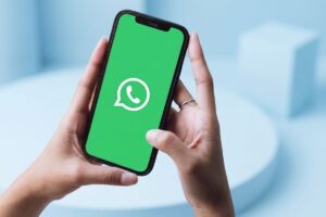 A imagem mostra um par de mãos segurando um smartphone com a tela ligada, exibindo o logotipo branco do WhatsApp sobre um fundo verde brilhante. O fundo da imagem é em tons claros de azul, com formas geométricas abstratas desfocadas, criando um ambiente minimalista e moderno. A foto é tirada de um ângulo superior, focando nas mãos e no dispositivo.