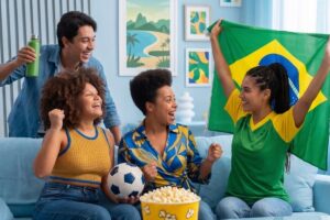 Quatro amigos reunidos em um sofá comemorando e assistindo a um jogo do Brasil, segurando uma bandeira e uma bola de futebol com um balde de pipoca na frente.