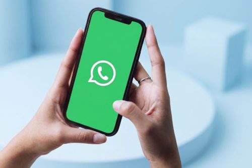 A imagem mostra um par de mãos segurando um smartphone com a tela ligada, exibindo o logotipo branco do WhatsApp sobre um fundo verde brilhante. O fundo da imagem é em tons claros de azul, com formas geométricas abstratas desfocadas, criando um ambiente minimalista e moderno. A foto é tirada de um ângulo superior, focando nas mãos e no dispositivo.