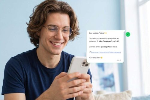 Um jovem com óculos dourados e uma camiseta azul-marinho sorri enquanto digita no celular, que tem uma capinha branca. Ele tem cabelos castanhos e ondulados.