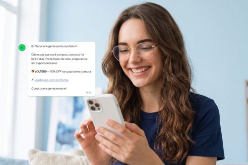 Uma jovem com óculos dourados e uma camiseta azul-marinho sorri enquanto digita no celular, que tem uma capinha branca. Ela tem cabelos castanhos longos e ondulados.