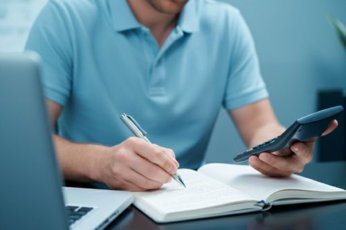 Homem calculando custos e escrevendo em caderno com calculadora ao lado do notebook