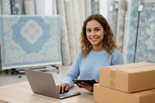 Mulher trabalhando em loja de tapetes com notebook e caixas de envio, representando vendas em marketplaces