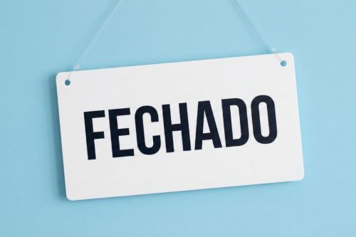 Uma placa retangular branca de PVC pendurada por um fio de nylon fino e transparente contra um fundo azul claro suave. A placa está levemente torta para a esquerda e traz a palavra "Fechado" escrita no centro com tipografia nítida e escura.